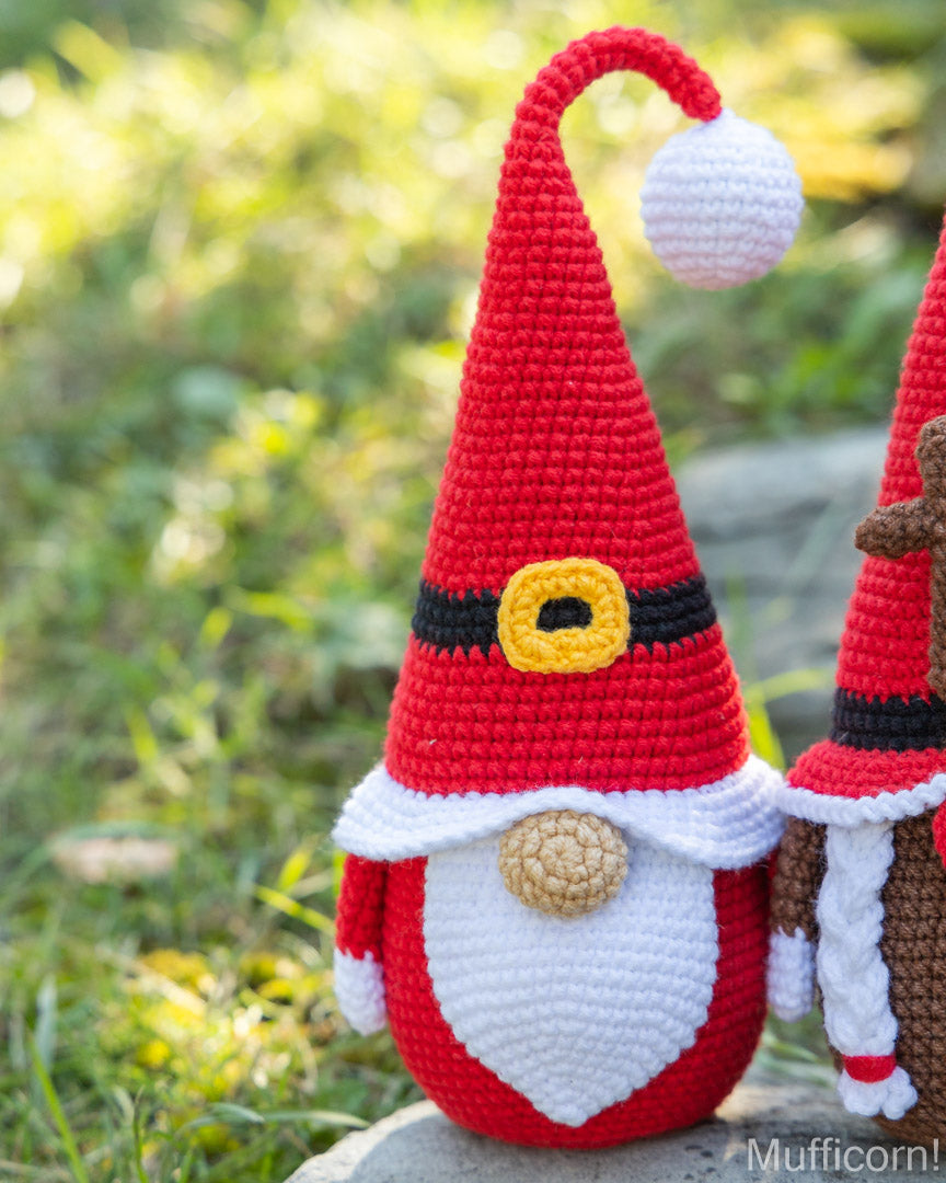 A crocheted Christmas gnome doll in red, white, and black with a yellow accent, standing outdoors on a stone surface with a blurred grassy background. Another similar gnome is partially visible on the right.